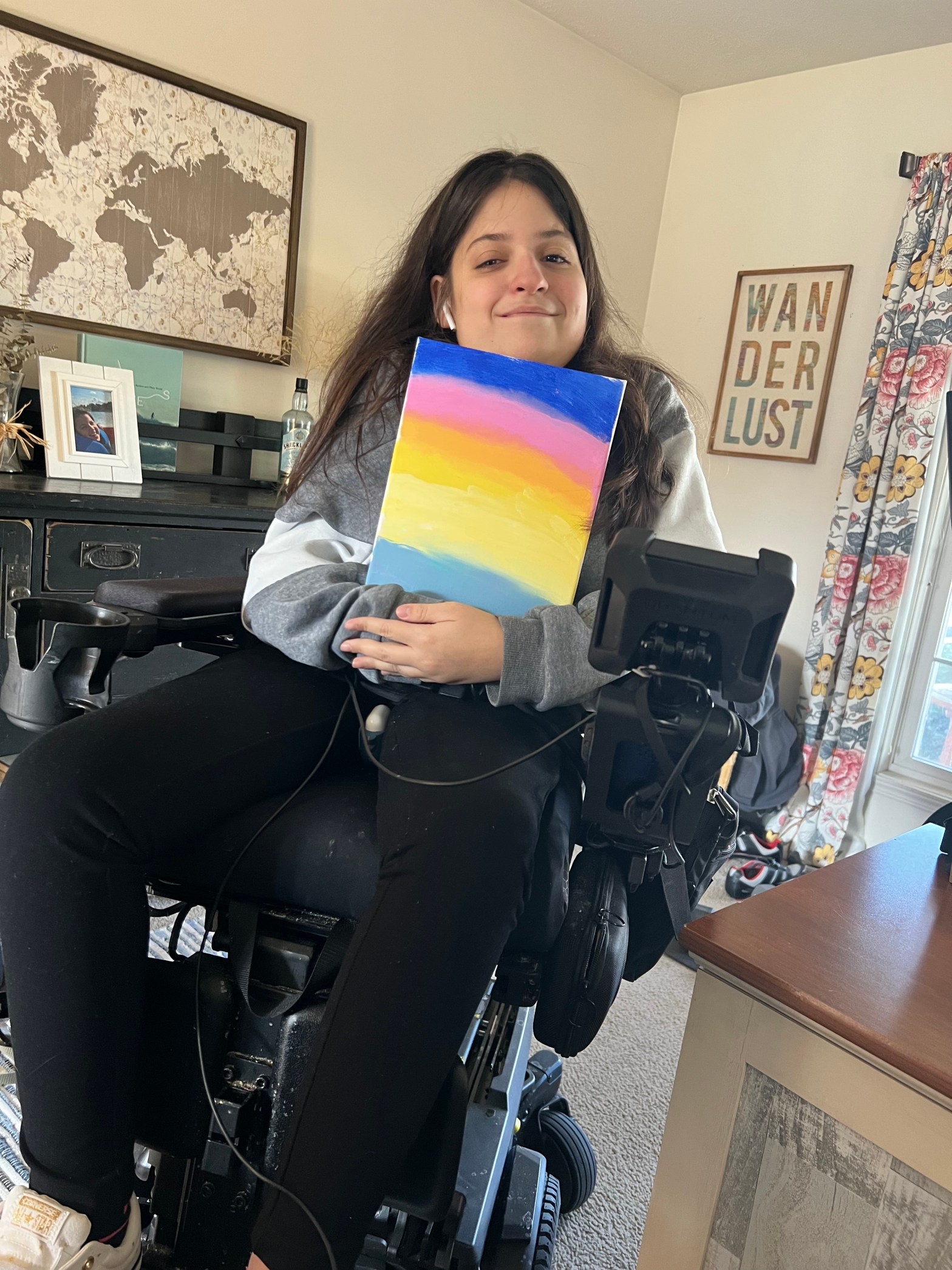 Image of Giana holding a rainbow painting in front of her. She is smiling and sitting in her Quantum Rehab power wheelchair with iLevel seat elevation technology.