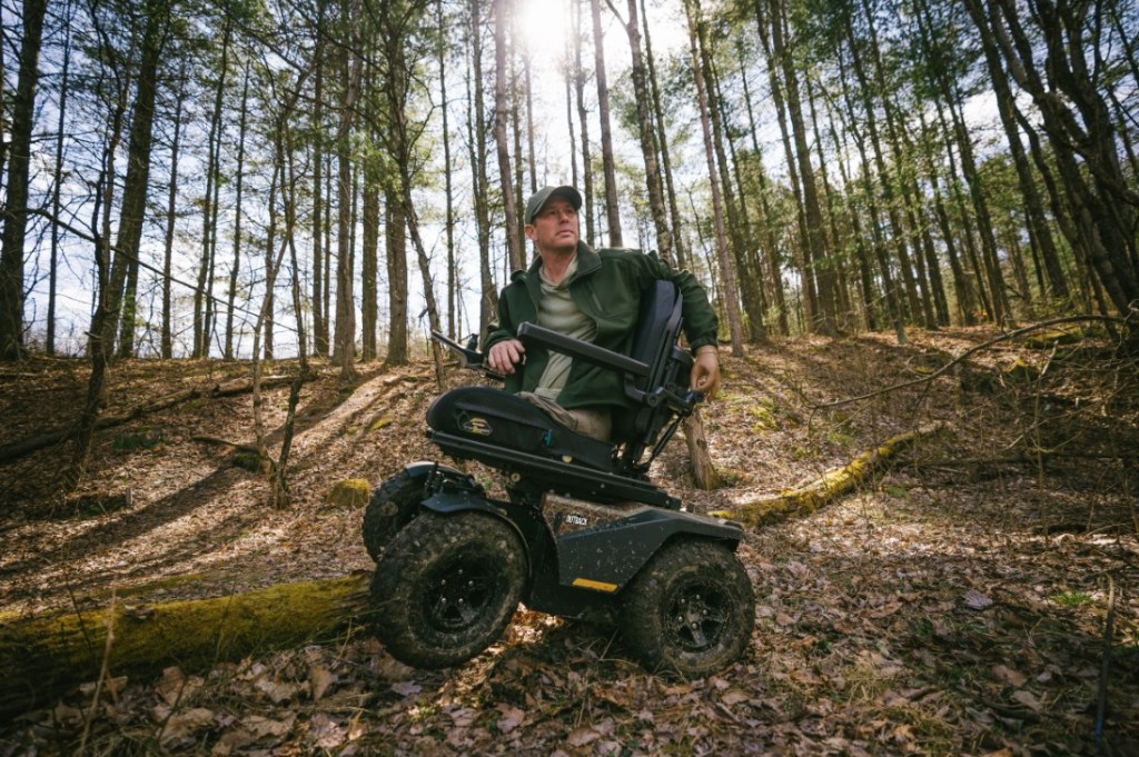 Photo of Bryan in the woods, he is seated in a Quantum Rehab Outback power wheelchair. The sun is shining through the trees and Bryan is looking toward the camera into the distance.